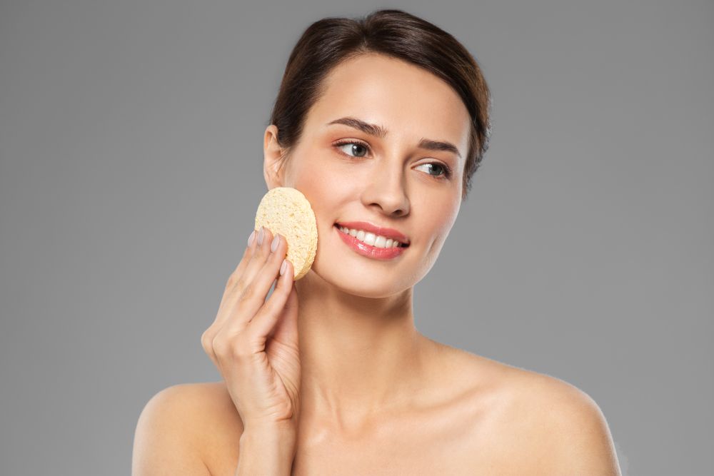 beauty, people and skincare concept - young woman cleaning face with exfoliating sponge over grey background. young woman cleaning face with exfoliating sponge