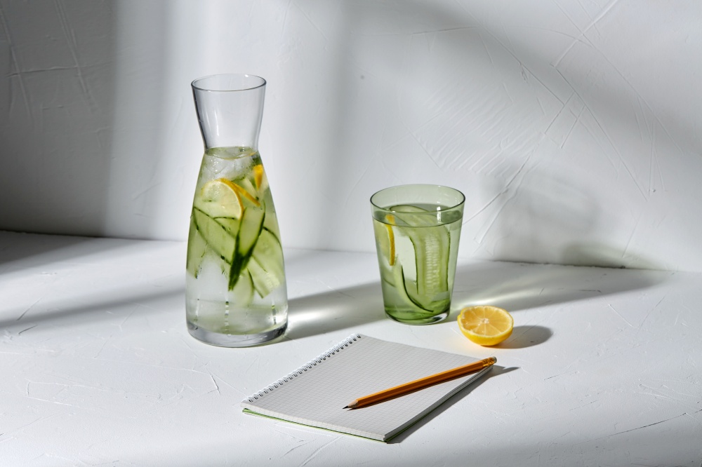 drink, detox and diet concept - glasses with fruit water with lemon and cucumber and notebook with pencil dropping shadows on white surface. water with lemon and cucumber and notebook