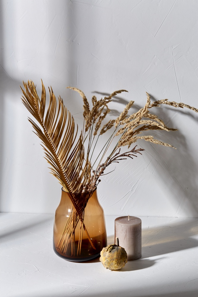 home improvement and decoration concept - still life of decorative dried flowers in brown glass vase, candle and pumpkin dropping shadows on white surface. dried flowers in glass vase candle and pumpkin