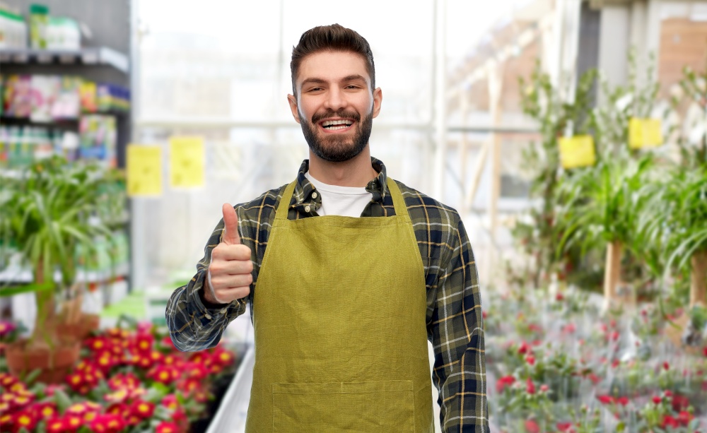 gardening, small business and sale concept - happy smiling male gardener or seller in apron showing thumbs up over flower shop background. happy gardener showing thumbs up at flower shop