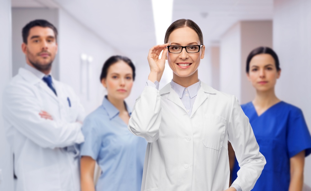 medicine, vision and healthcare concept - happy smiling female doctor in glasses and white coat over colleagues at hospital on background. smiling female doctor with colleagues at hospital