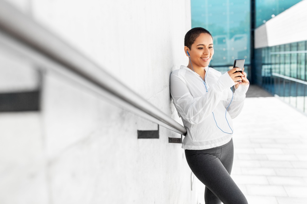 fitness, sport and technology concept - happy smiling young african american woman with earphones listening to music on smartphone outdoors. african american woman with earphones and phone