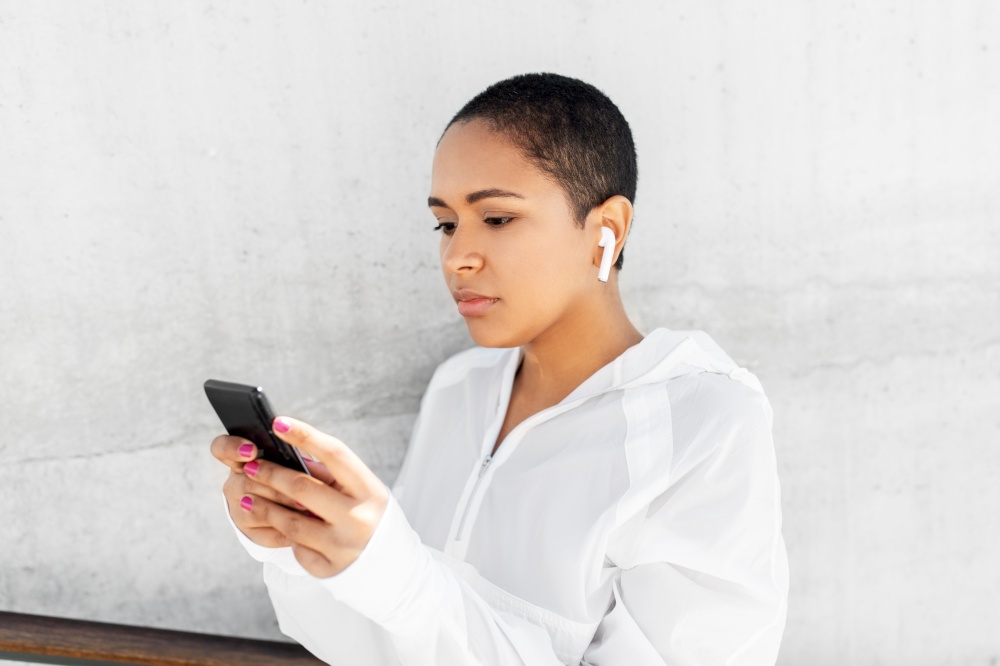 fitness, sport and technology concept - young african american woman with wireless earphones listening to music on smartphone outdoors. african american woman with earphones and phone