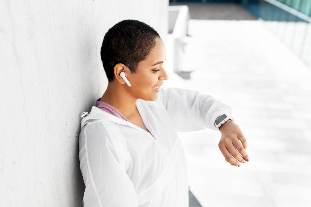 fitness, sport and technology concept - young african american woman with earphones listening to music on smart watch outdoors. african woman with earphones and smart watch