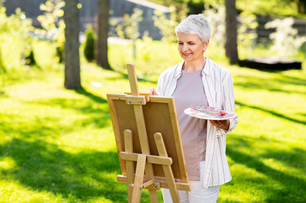 creativity, art and hobby concept - senior woman with easel and color palette painting outdoors. senior woman with easel painting outdoors