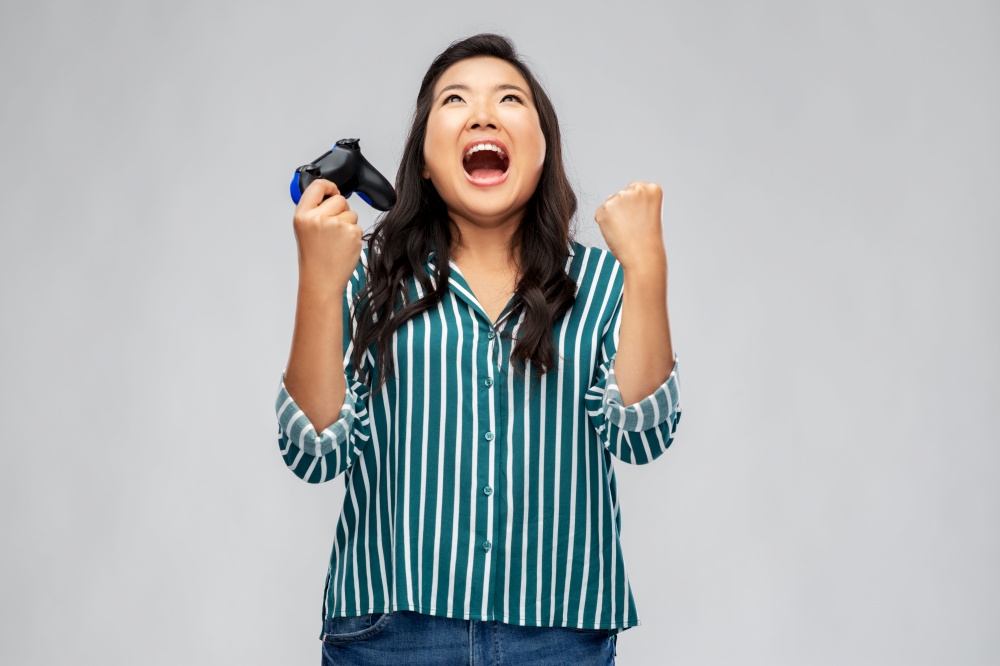 people, leisure and gaming concept - happy asian young woman playing video game with gamepad celebrating success over grey background. happy asian woman playing video game with gamepad
