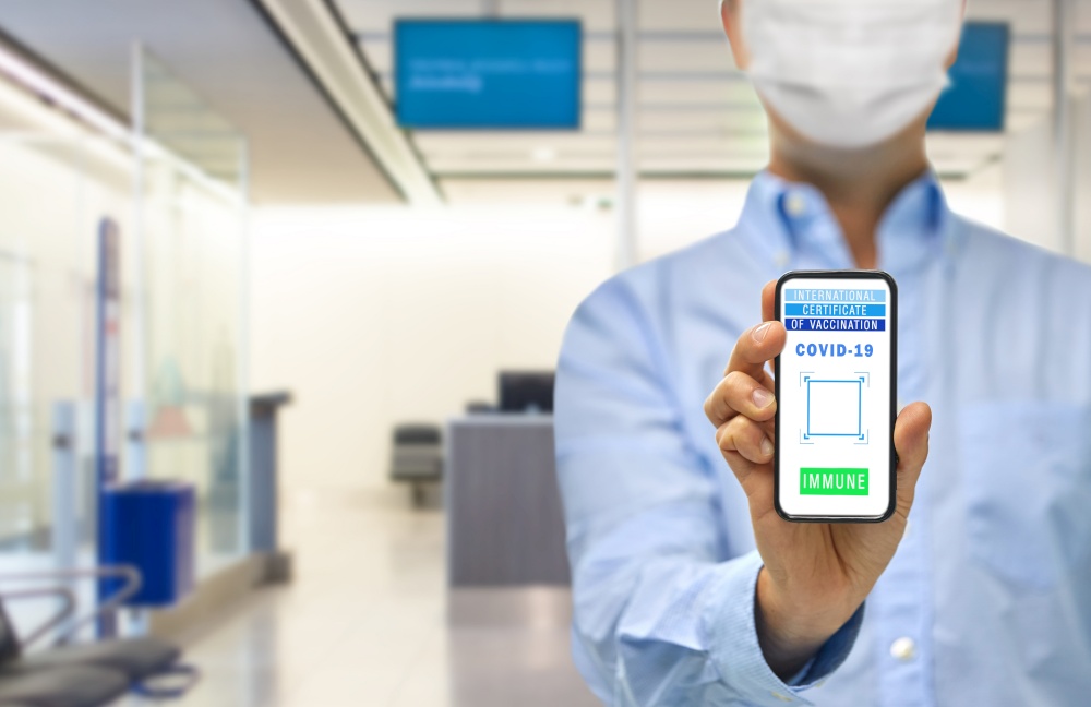 safe travel, technology and health care concept - close up of man holding and showing smartphone with international certificate of vaccination on screen over airport background. hand with virtual immunity passport on smartphone