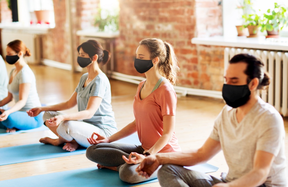 fitness, yoga and healthy lifestyle concept - group of people wearing face protective black masks for protection from virus disease meditating in lotus pose at studio. group of people in masks doing yoga at studio