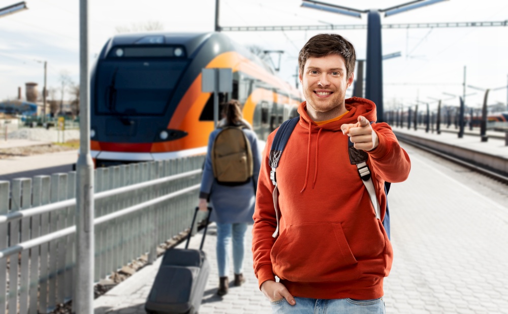 travel, tourism and people concept - smiling young man with backpack pointing finger to camera traveling by train over railway station in city of tallinn, estonia on background. smiling man with backpack traveling by train