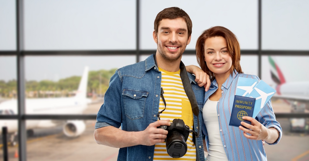 safe travel, tourism and vacation concept - happy couple with air tickets, immunity passport and camera over airport background. couple with tickets, immunity passport and camera