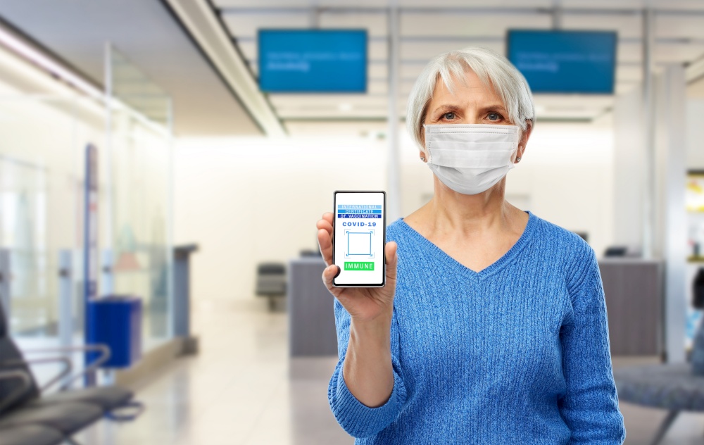 safe travel, technology and health care concept - senior woman in mask holding and showing smartphone with international certificate of vaccination on screen over airport background. old woman with certificate of vaccination on phone