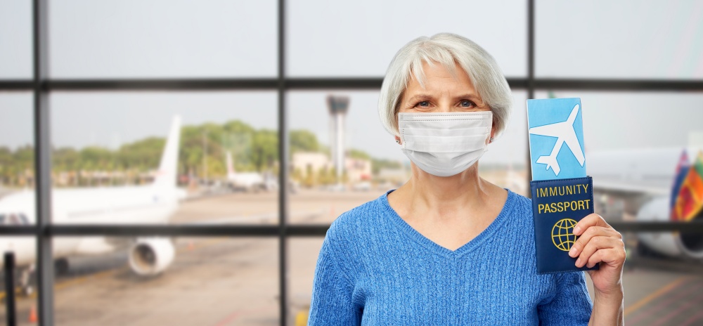 tourism, safe travel and vacation concept - senior woman in mask with immunity passport and airplane ticket over airport background. old woman in mask with immune passport and ticket