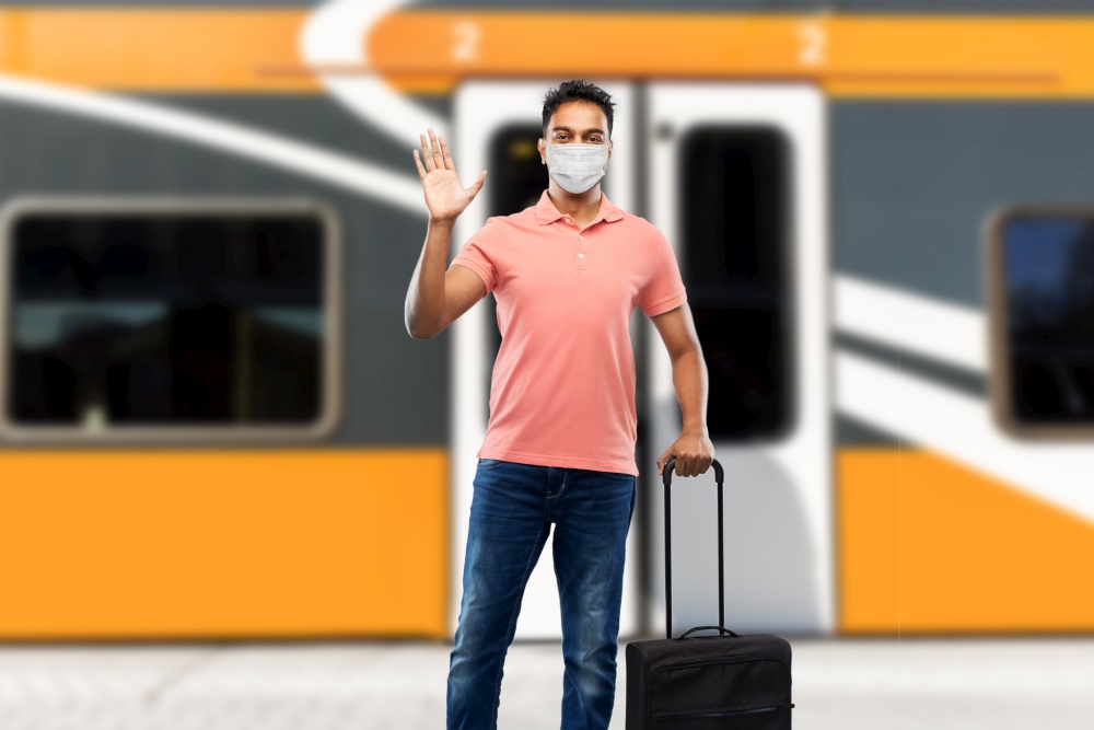 tourism, vacation and health care concept - indian man in face protective mask for protection from virus disease with travel bag waving hand over train on railway station on background. indian man in mask with travel bag waving hand