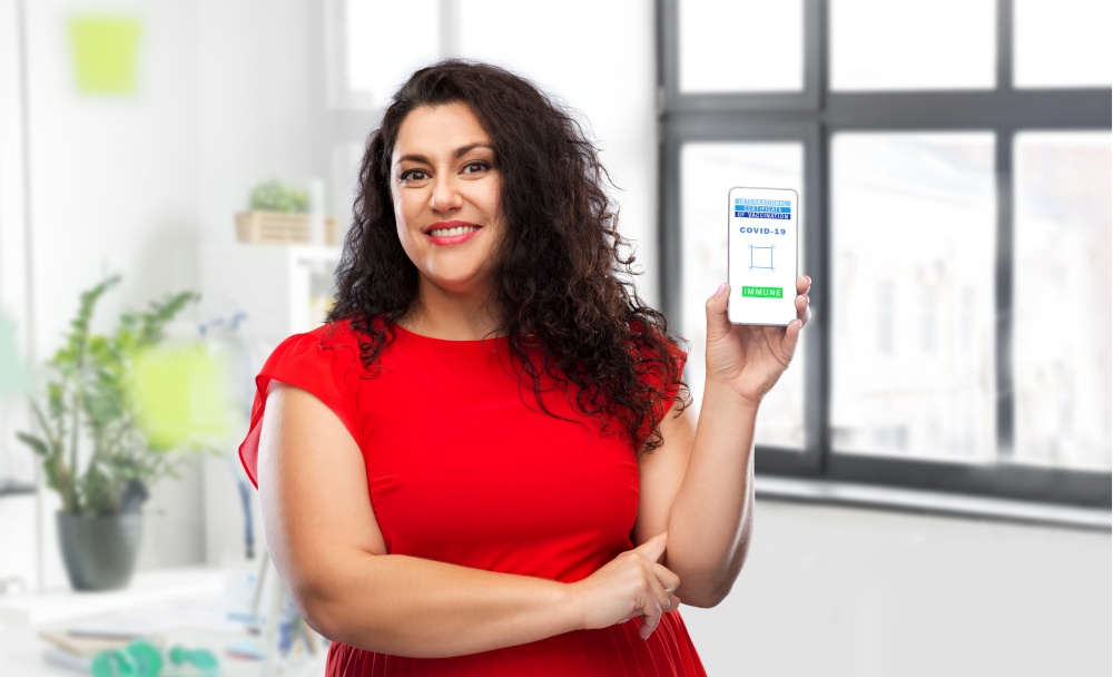 technology and health care concept - close up of happy smiling woman holding and showing smartphone with international certificate of vaccination on screen over office background. woman with certificate of vaccination on phone