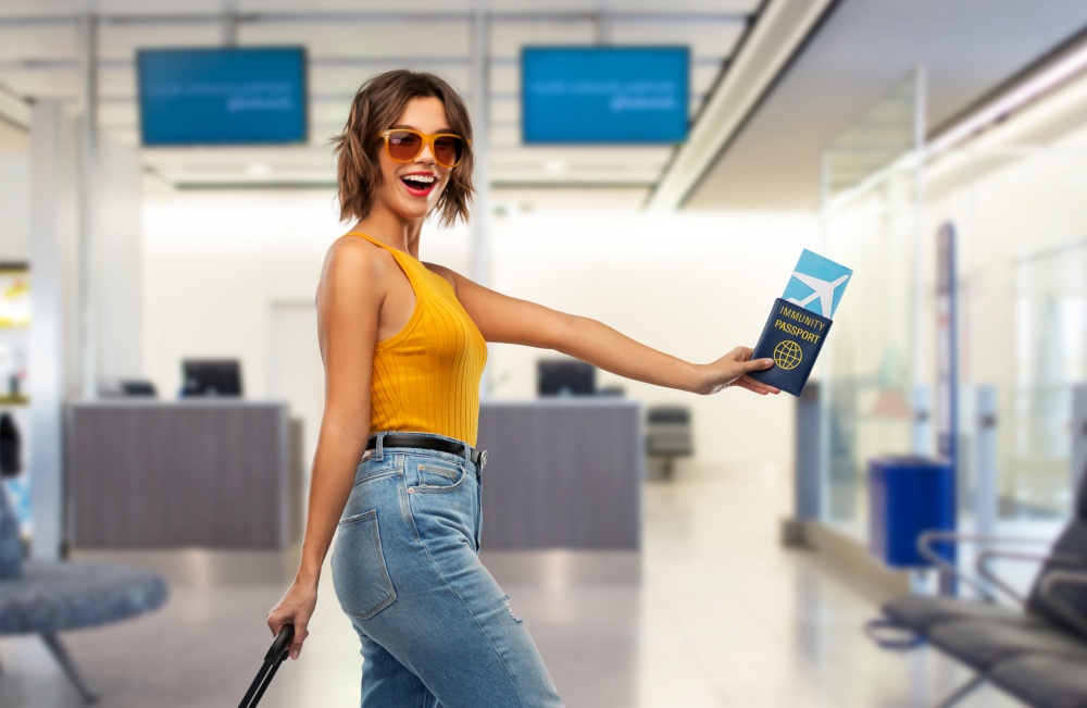 safe travel, tourism and health care concept - happy laughing young woman with air ticket, immunity passport and bag walking over airport background. happy woman with air ticket and immunity passport