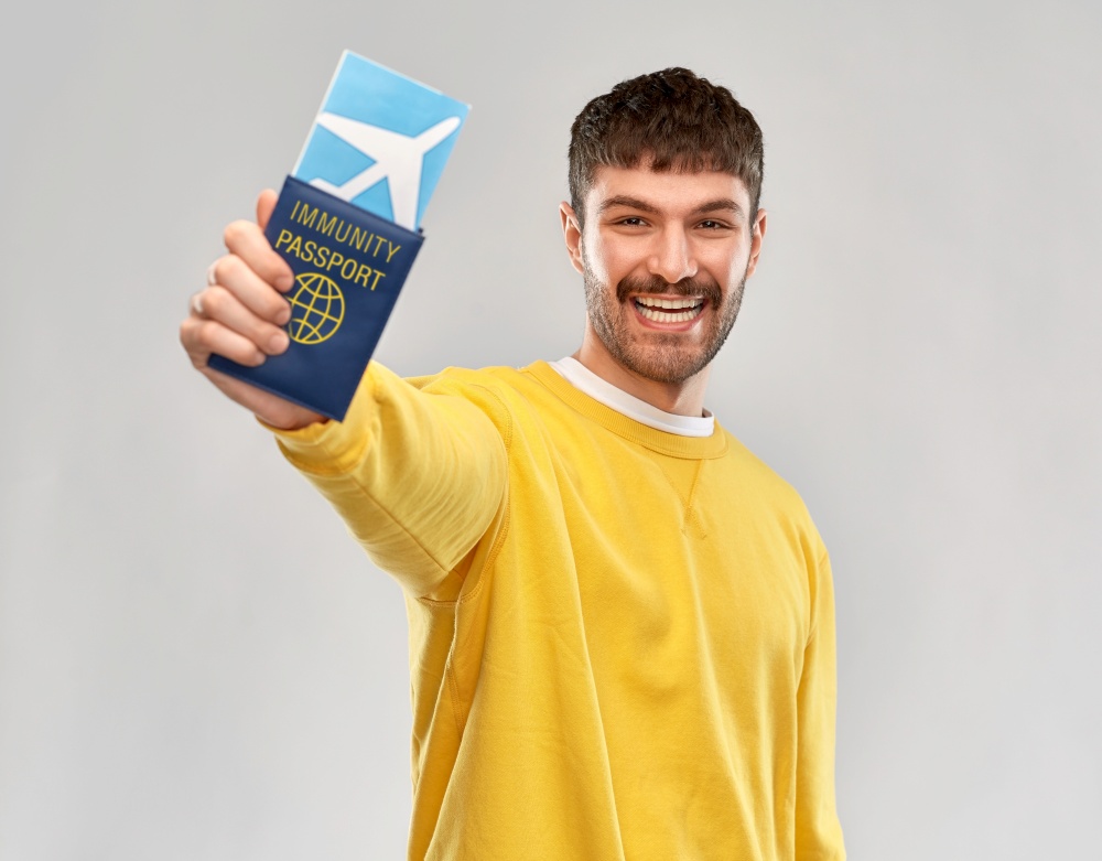 safe travel, tourism and health care concept - smiling young man in yellow sweatshirt with air ticket and immunity passport over grey background. smiling man with air ticket and immunity passport