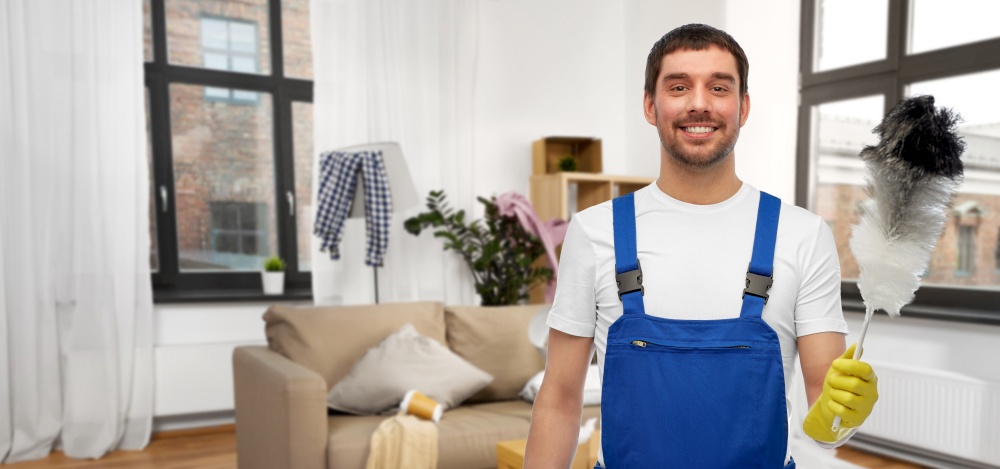 profession, cleaning service and people concept - happy smiling male worker or cleaner in overall and gloves with duster over home living room background. happy male cleaner with duster cleaning home room