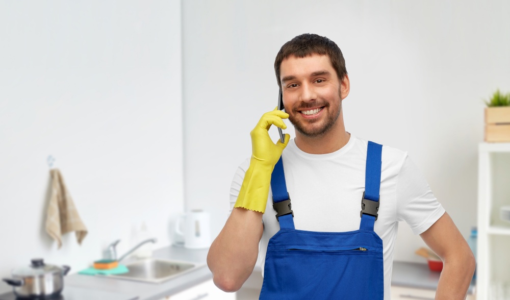 profession, cleaning service and plumbing concept - happy smiling male worker, plumber or cleaner in overall and gloves calling on smartphone over home room background. male worker or cleaner calling on phone at kitchen