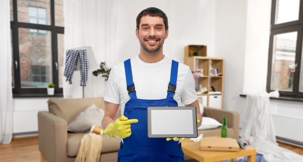 profession, cleaning service and people concept - happy smiling male worker or cleaner in overall and gloves showing tablet pc computer over home room background. male worker or cleaner showing tablet pc at home