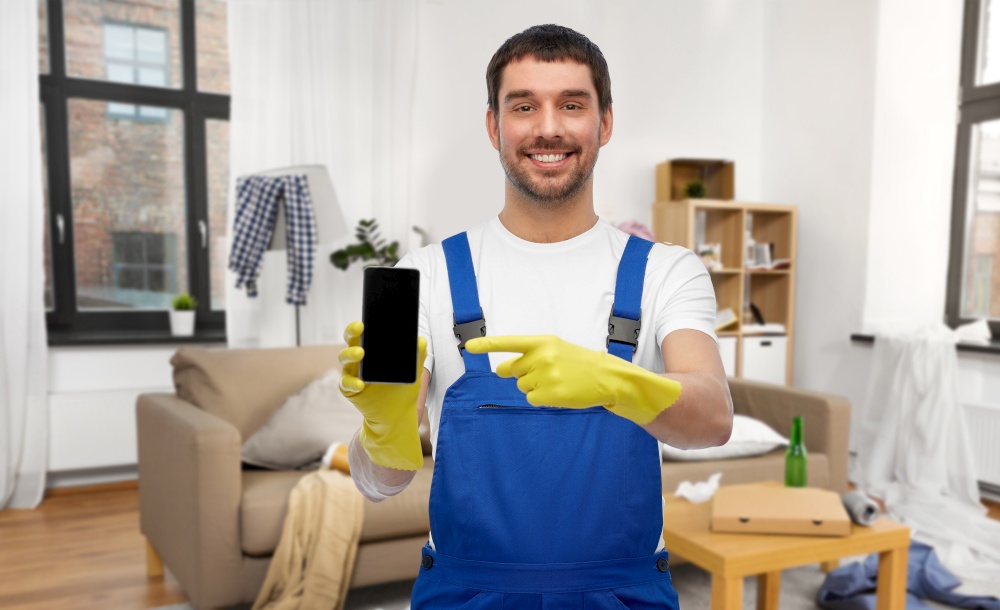 profession, cleaning service and people concept - happy smiling male worker or cleaner in overall and gloves showing smartphone over home room background. male worker or cleaner showing smartphone at home