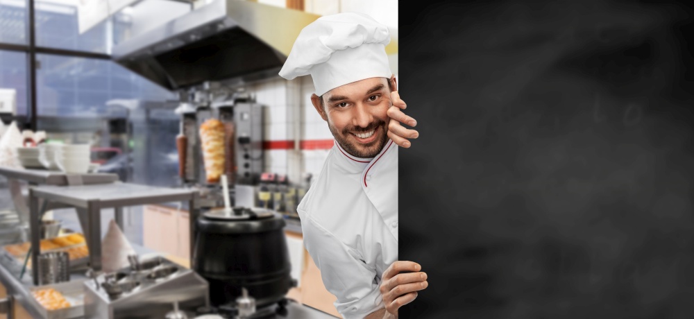 cooking, culinary and people concept - happy smiling male chef in toque with big black chalkboard over restaurant kitchen background. happy smiling male chef with big black chalkboard