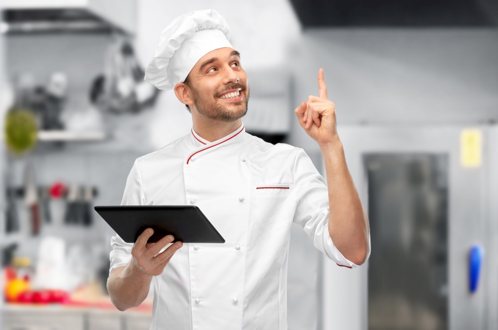 cooking, culinary and people concept - happy smiling male chef in toque with tablet pc computer pointing finger up over restaurant kitchen background. male chef with tablet computer pointing finger up