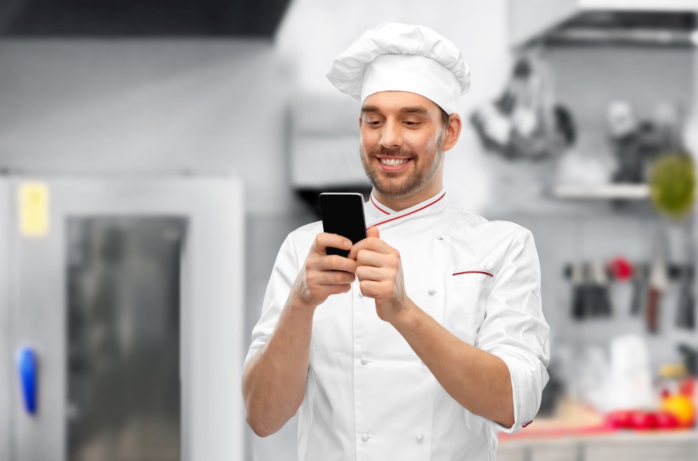 cooking, culinary and people concept - happy smiling male chef in toque with smartphone over restaurant kitchen background. happy smiling male chef with smartphone at kitchen