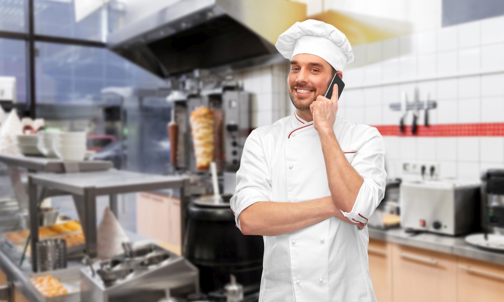cooking, culinary and people concept - happy smiling male chef in toque and jacket calling on smartphone over restaurant kitchen background. happy smiling male chef calling on smartphone