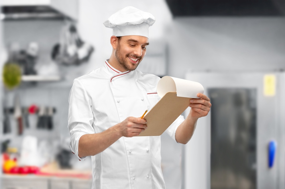 cooking, culinary and people concept - happy smiling male chef with clipboard and pencil over restaurant kitchen background. happy smiling male chef with clipboard at kitchen