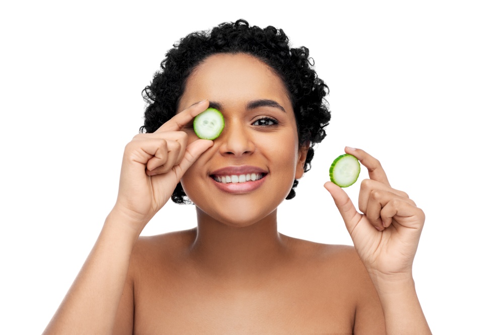 beauty, skin care and detox concept - beautiful african american woman making eye mask of cucumbers over white background. happy african woman making eye mask of cucumbers