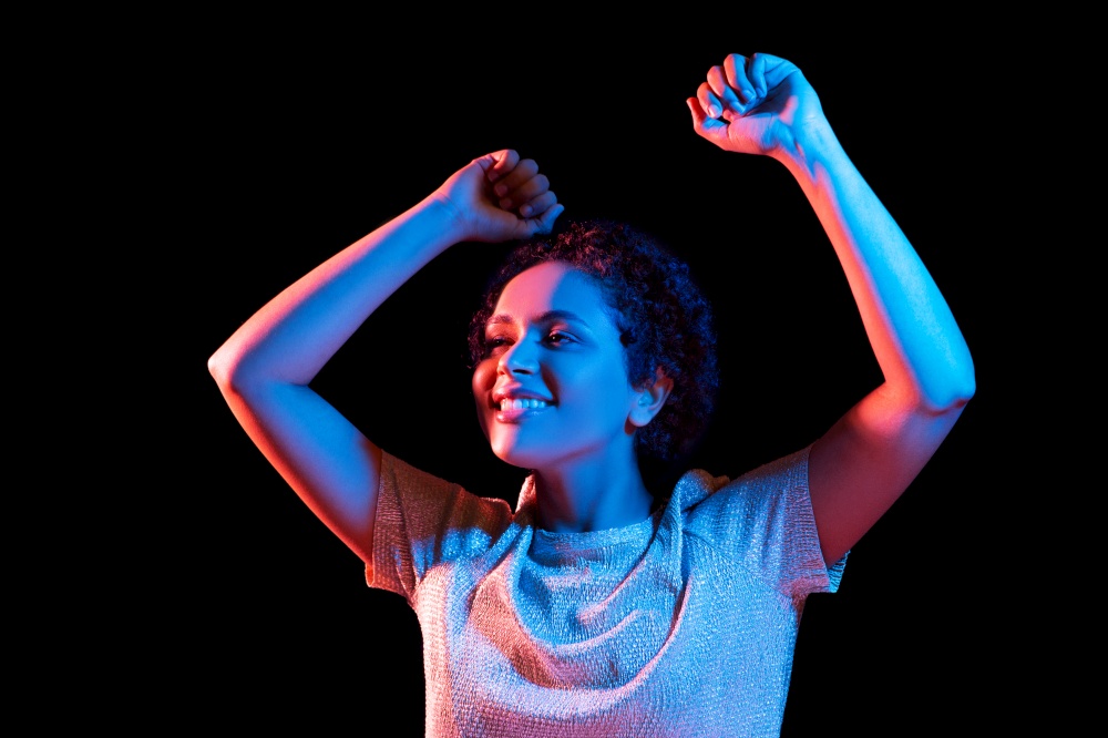 leisure, clubbing and nightlife concept - smiling young african american woman dancing over ultraviolet neon lights on black background. african american woman dancing over neon lights