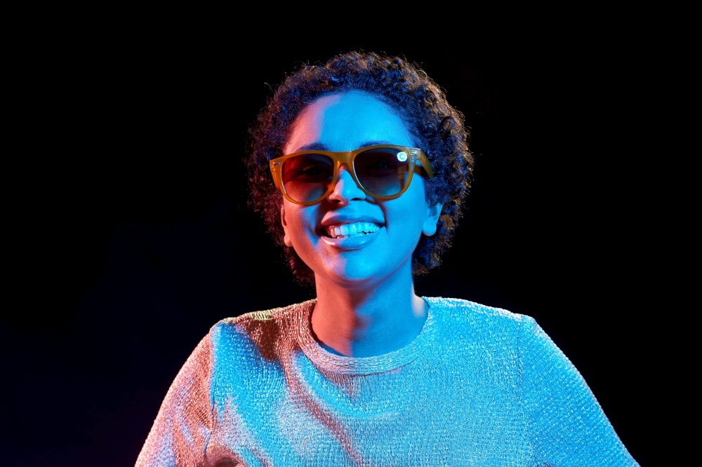 leisure, clubbing and nightlife concept - smiling young african american woman in sunglasses over ultraviolet neon lights on black background. african woman in sunglasses over neon lights