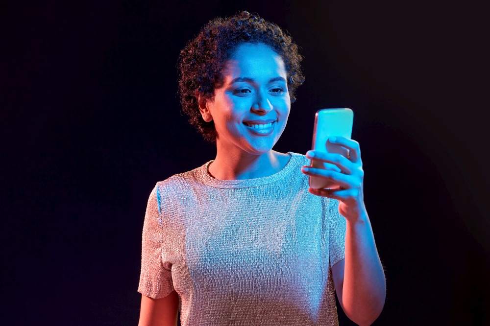 nightlife, technology and people concept - happy young african american woman with smartphone in neon lights over black background. african woman with smartphone in neon lights