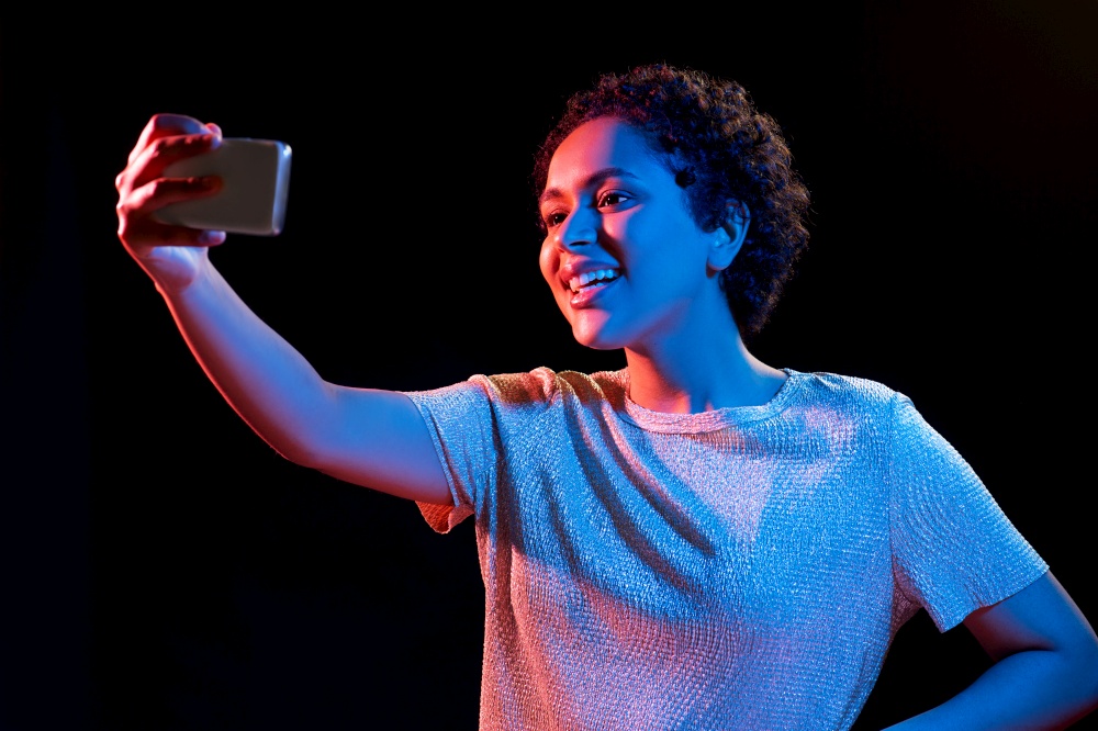 nightlife, technology and people concept - happy young african american woman taking selfie with smartphone in neon lights over black background. woman taking selfie with smartphone in neon lights