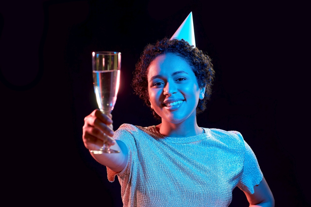 birthday, celebration and nightlife concept - happy smiling young african american woman in party cap with glass of champagne over ultraviolet neon lights on black background. african woman in party cap with glass of champagne