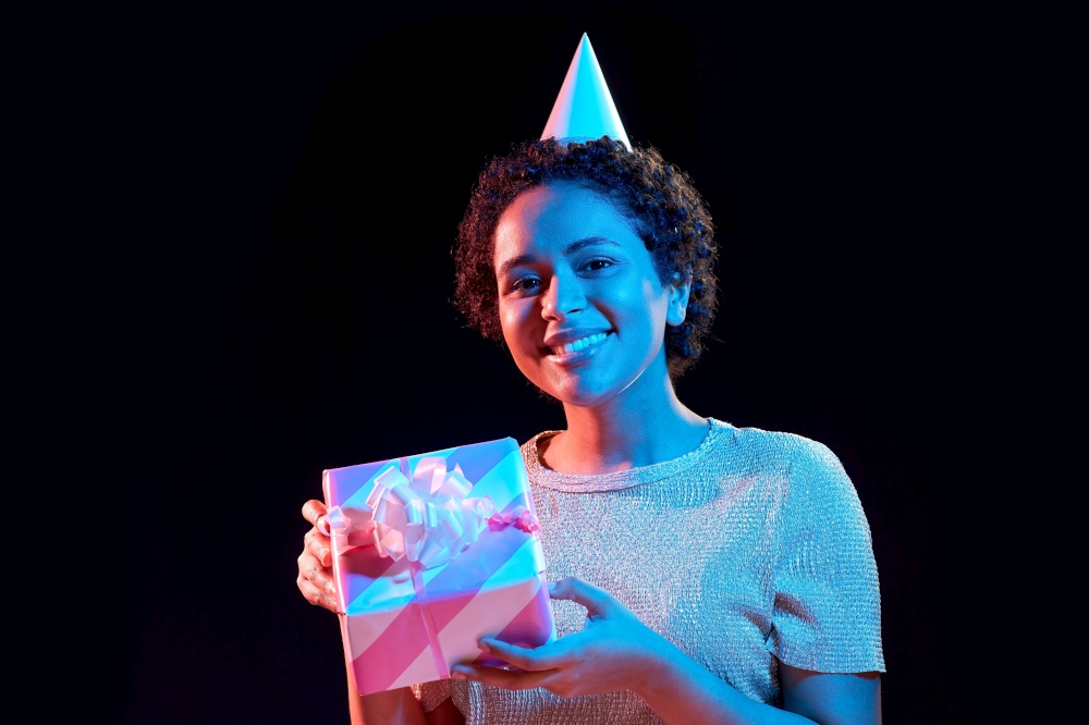 birthday, celebration and nightlife concept - happy smiling young african american woman in party cap with gift box over ultraviolet neon lights on black background. african woman in party cap with gift box on black
