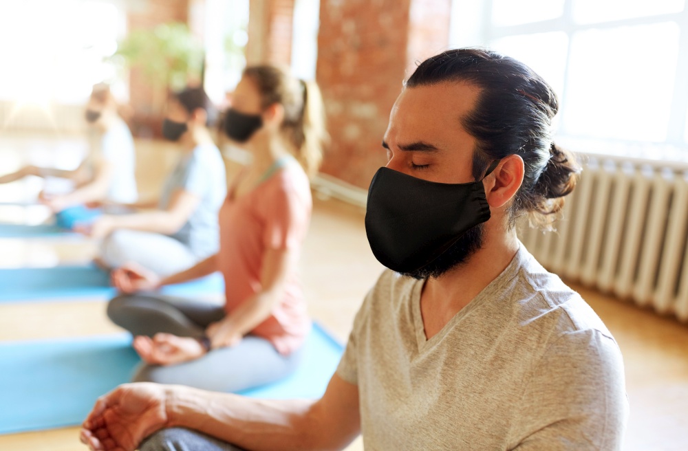 fitness, yoga and health care concept - man with group of people wearing black face protective masks for protection from virus disease meditating in lotus pose at studio. group of people in masks doing yoga at studio