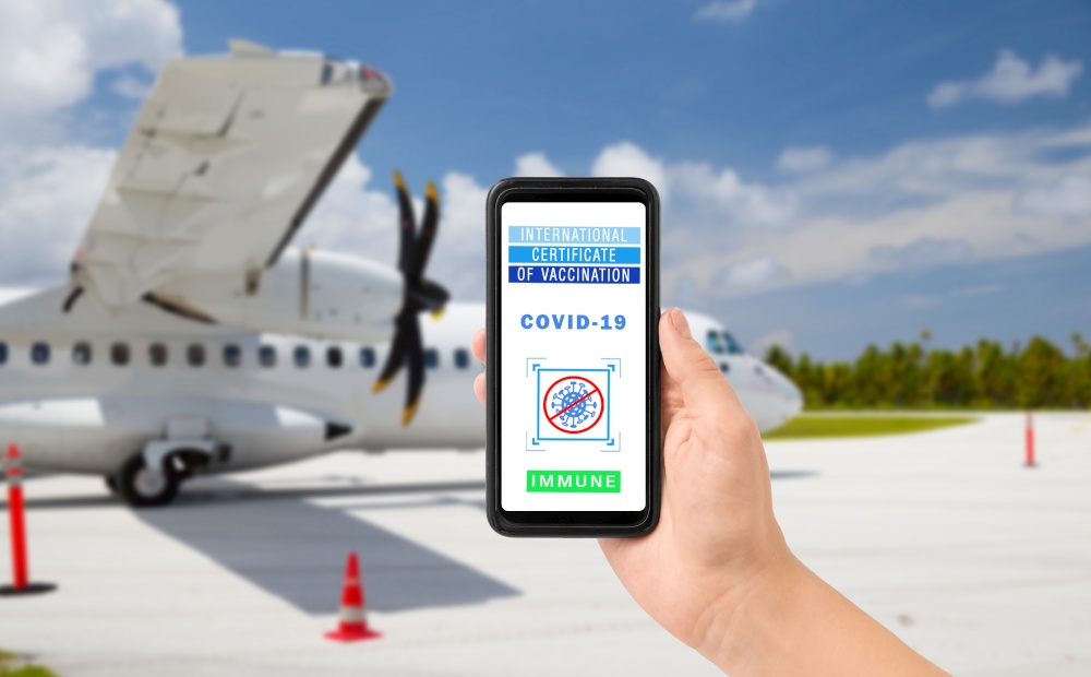 safe travel, technology and health care concept - close up of hand holding and showing smartphone with international certificate of vaccination on screen over plane at airport background. hand holding phone with certificate of vaccination