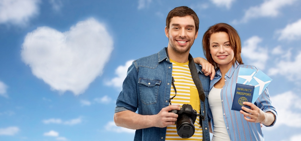 safe travel, tourism and vacation concept - happy couple with air tickets, immunity passport and camera over blue sky and clouds background. couple with tickets, immunity passport and camera