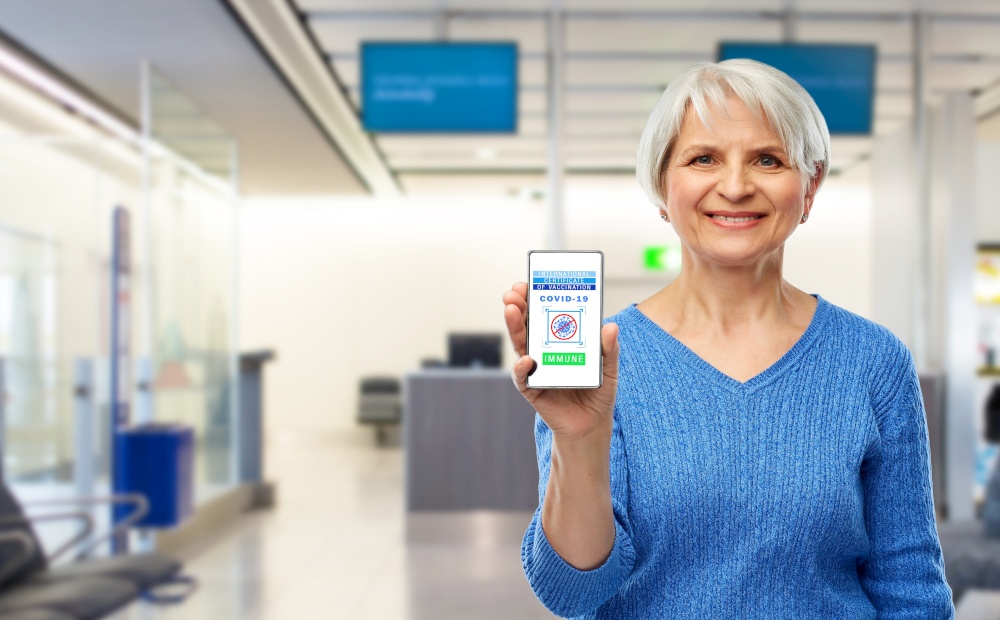safe travel, technology and health care concept - happy smiling senior woman holding and showing smartphone with international certificate of vaccination on screen over airport background. old woman with certificate of vaccination on phone