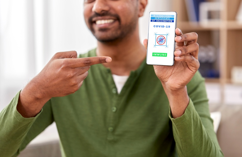 technology and health care concept - close up of smiling indian man holding and showing smartphone with international certificate of vaccination on screen over home background. man with certificate of vaccination on smartphone