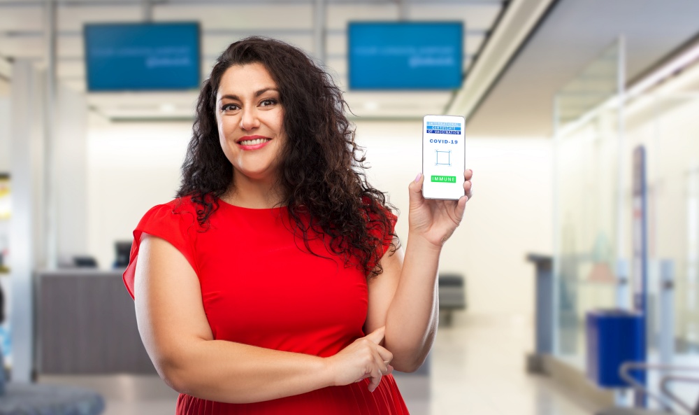 safe travel, technology and health care concept - close up of happy smiling woman holding and showing smartphone with international certificate of vaccination on screen over airport background. woman with certificate of vaccination on phone