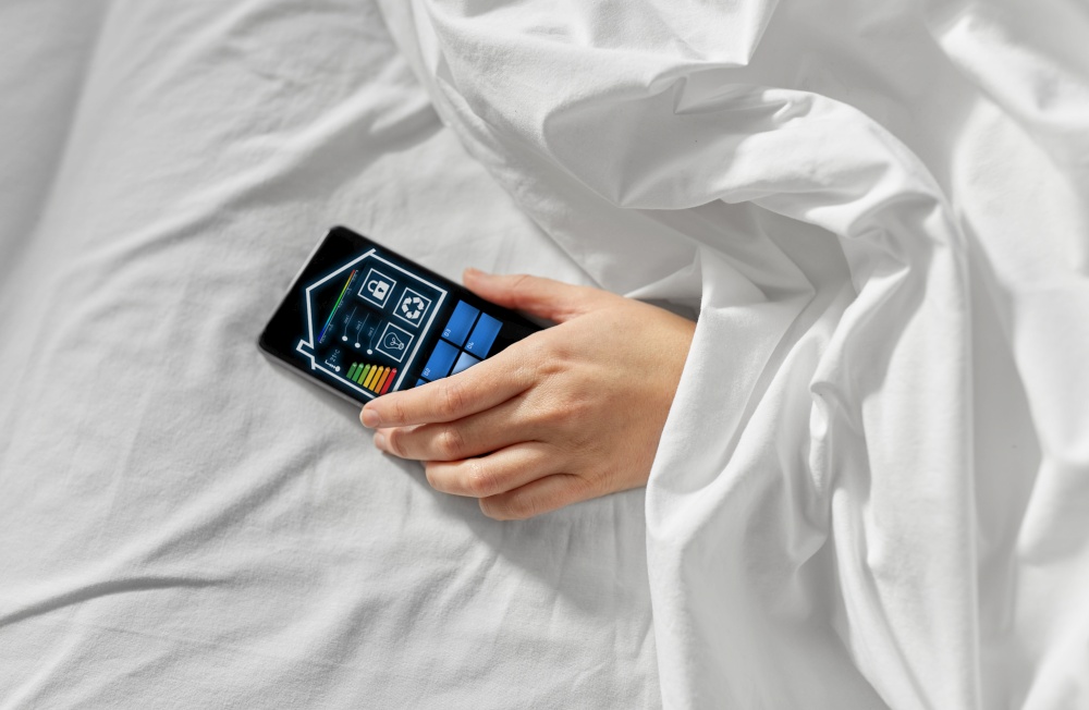 technology, efficiency and people concept - hand of young woman lying in bed with smart home icons on smartphone screen at bedroom. hand in bed with smart home icons on smartphone