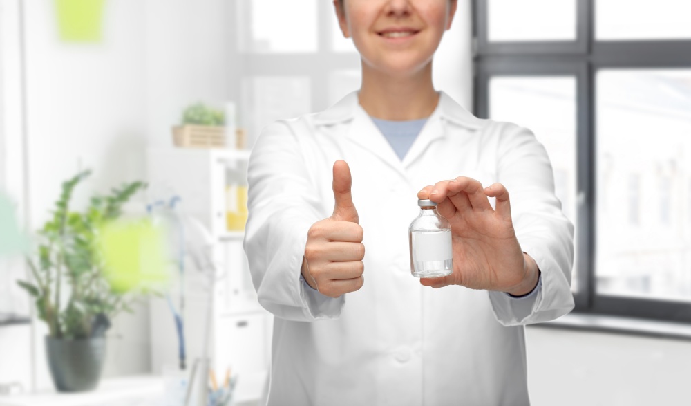 medicine, vaccination and healthcare concept - close up of happy smiling female doctor or nurse with drug in bottle showing thumbs up over medical office at hospital background. doctor with medicine showing thumbs up at hospital