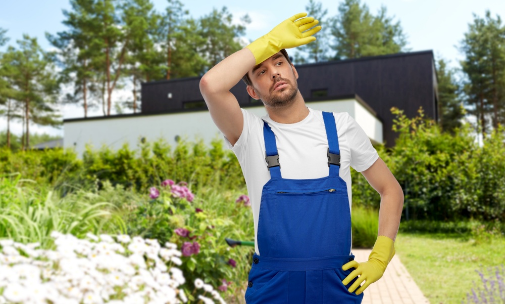 profession, cleaning service and people concept - tired male worker or cleaner in overall and gloves over summer garden background. tired male worker or cleaner in gloves at garden
