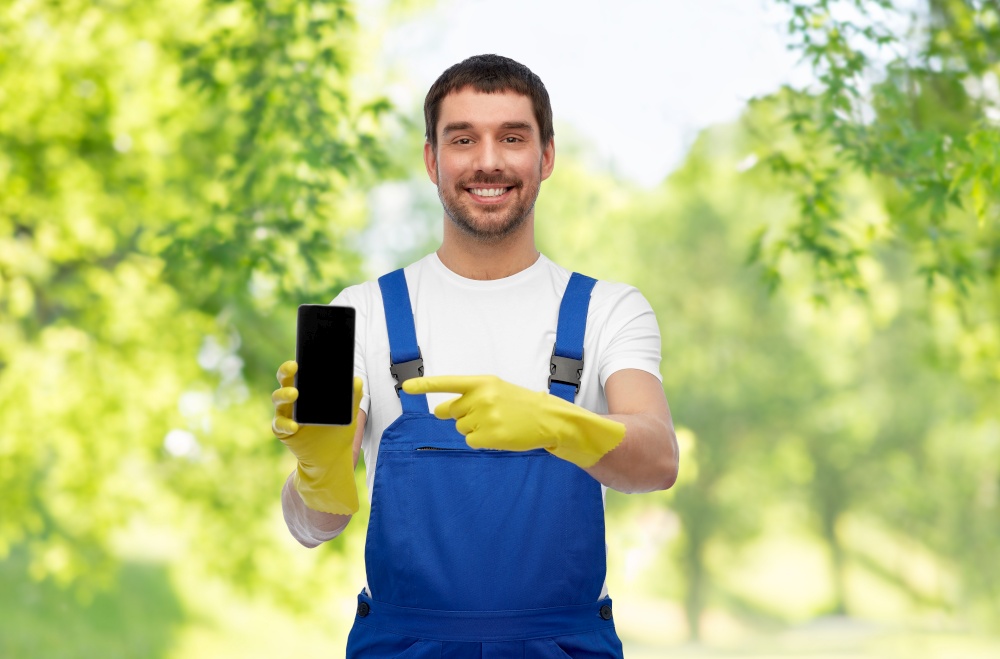profession, cleaning service and people concept - happy smiling male worker or cleaner in overall and gloves showing smartphone over green natural background. happy male worker or cleaner showing smartphone
