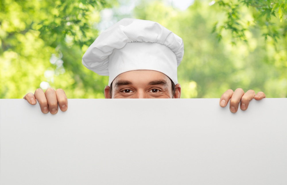cooking, culinary and people concept - male chef in toque hiding behind big white board over green natural background. male chef hiding behind big white board