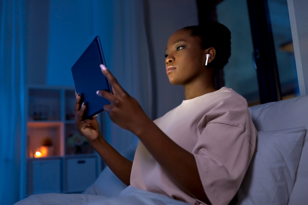 technology, internet and people concept - young african american woman with tablet pc computer and wireless earphones lying in bed at home at night. woman with tablet pc in earphones in bed at night