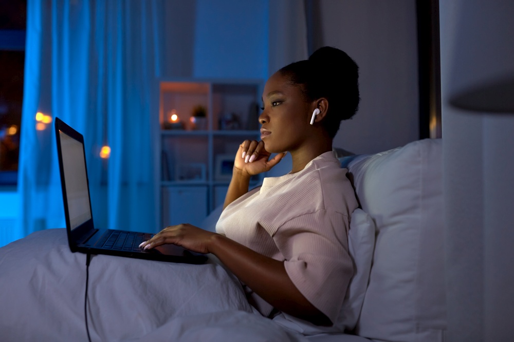 technology, internet, communication and people concept - young african american woman with laptop computer and earphones in bed at home at night. woman with laptop and earphones in bed at night