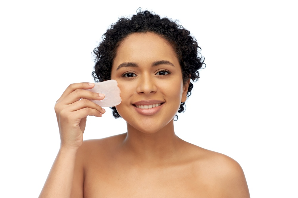 beauty and people concept - portrait of happy smiling young african american woman massaging her face with gua sha facial tool over white background. happy woman massaging her face with gua sha tool